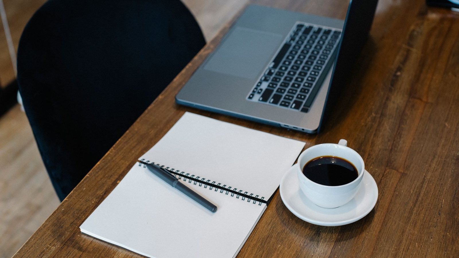 An open laptop and a spiral notebook with blank pages on a wooden table, beside a cup of coffee and a pen.