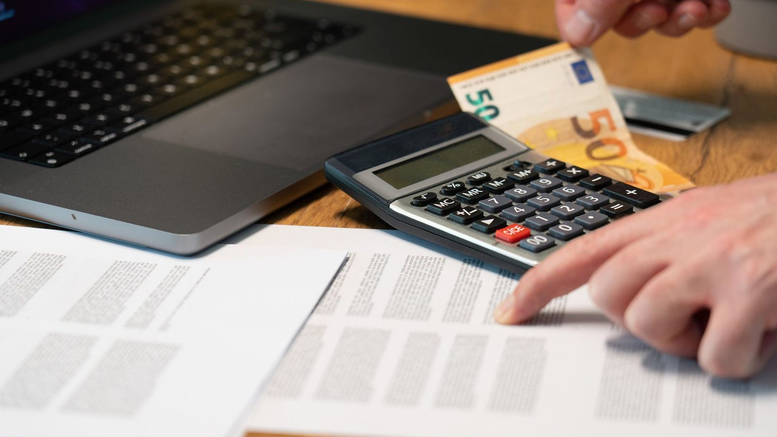 A hand working a calculator beside euro banknotes, a laptop and printed figures, the setting in which Belgian state costs get added up.